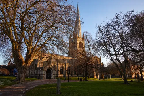 Church of St Mary and All Saints winter morning The image shows an architectural photograph of the Church of St Mary and All Saints in Chesterfield, taken on a winter morning. The church is notable for its historic spire and is surrounded by leafless trees, indicating the season. The early morning sunlight casts a golden hue onto the stone façade, highlighting the church's details and the surrounding grounds. This view captures the tranquility of the Chesterfield landmark, with its prominent spire rising above the landscape in the quietness of a winter morning.